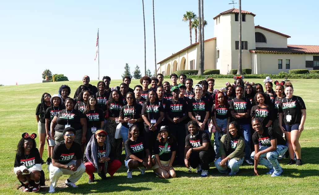 Large group of student participants wearing Black RISE shirts standing on the grass in front of Cal Poly Pomona's old horse stables